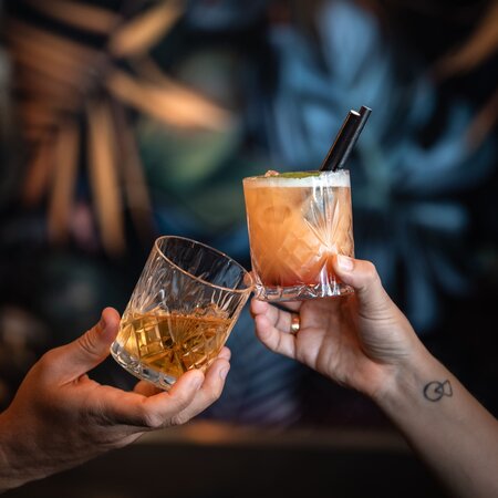 Zwei Hände stoßen mit einem Glas Whiskey auf Eis und einem farbigen Cocktail mit Strohhalmen an