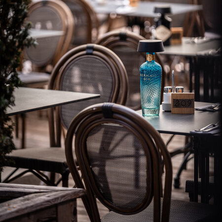 Leere geflochtene Stühle und Tische auf einer Restaurant-Terrasse mit einer blauen Ginflaschen-Lampe